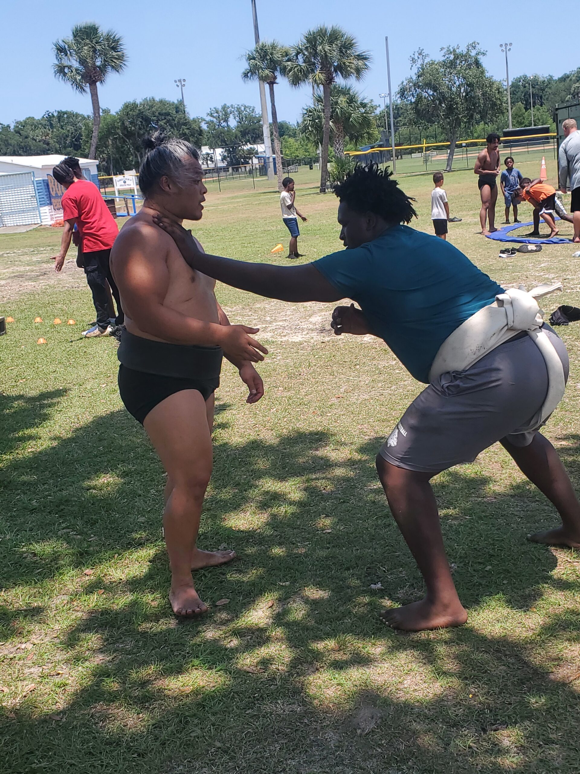 Two men practicing sumo wrestling outdoors on a sunny day, surrounded by onlookers They are in an athletic stance, engaged in a grapple, with palm trees and a field in the background