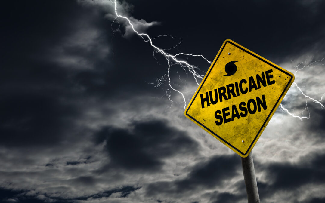 A yellow warning sign with the words "Hurricane Season" and a hurricane symbol stands against a dark, stormy sky with lightning in the background