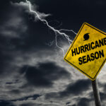 A yellow warning sign with the words "Hurricane Season" and a hurricane symbol stands against a dark, stormy sky with lightning in the background