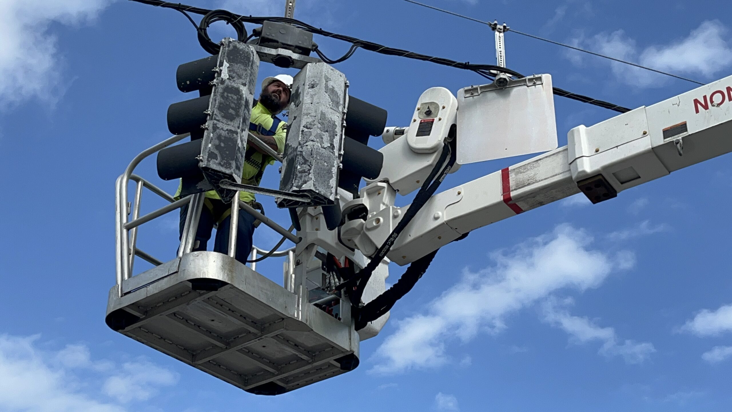 A worker in a cherry picker truck repairs a set of traffic lights The person is wearing a safety vest and helmet, positioned high above the street against a backdrop of blue sky and clouds