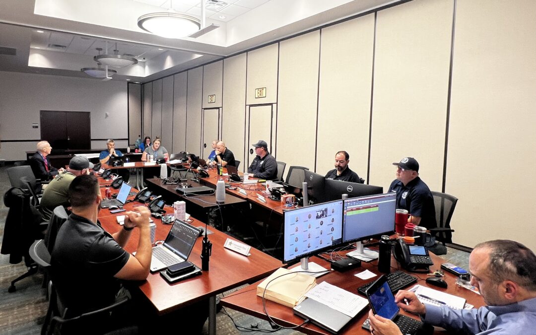 A group of people sitting at a large conference table with computers, laptops, and various equipment They appear to be engaged in a meeting or discussion in a well lit, modern office space