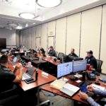 A group of people sitting at a large conference table with computers, laptops, and various equipment They appear to be engaged in a meeting or discussion in a well lit, modern office space