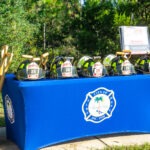 A display table with firefighting helmets and axes, marked with the number , under an American flag The table is covered with a blue cloth featuring the City of Palm Coast Fire Department emblem Trees and a sign are in the background