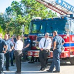 A group of firefighters, both men and women, in uniform stand and talk in front of a large red fire truck labeled "Palm Coast " They are near a green, wooded area on a sunny day One firefighter leans against the truck