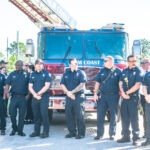 A group of firefighters wearing navy uniforms stands in front of a red fire truck with "Palm Coast" written on it They are outdoors, under a clear sky with greenery in the background