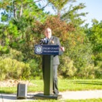 A person stands behind a podium with a "Palm Coast Fire Department" logo, speaking outdoors They are surrounded by greenery and trees, with a small speaker on the ground nearby The scene is bright and sunny