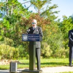 A person in a suit and sunglasses speaks at a podium labeled "Palm Coast Fire Department" outdoors There is a microphone, and greenery like trees and grass in the background under a clear sky