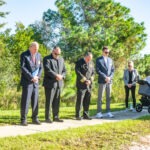 A group of people, including officials in suits and a person with a stroller, stand solemnly on a sunny pathway surrounded by greenery They are bowing their heads, possibly in remembrance or prayer