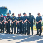 A group of uniformed firefighters stands solemnly in front of a fire truck, bowing their heads in a moment of silence The truck displays the American flag on the front Trees are visible in the background under a clear blue sky