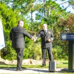 Two men in formal uniforms shake hands near a podium with a "Palm Coast Fire Department" sign A white flag and green trees are in the background, and a pile of dirt is visible to the side