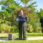 A person in a Palm Coast Fire Department uniform speaks at a podium outdoors, surrounded by trees and greenery A speaker is placed on the ground nearby