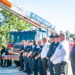 A lineup of firefighters and officials stands in front of a Palm Coast fire truck with an extended ladder They are outdoors on a sunny day, wearing uniforms and badges Trees and clear blue sky are visible in the background