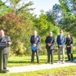 A group of six people stand outdoors on a sunny day One person is speaking at a podium with “Palm Coast Fire Department” written on it The others stand in a line, with trees and greenery in the background