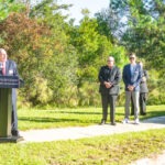 A man speaks at a podium with the Palm Coast Fire Department logo, outdoors Four people, including a man in sunglasses and a person with a stroller, stand nearby Trees and greenery are in the background on a sunny day