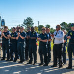 A group of uniformed firefighters and emergency personnel stand in a line outdoors, clapping A fire truck is partially visible in the background The sky is clear and sunny, and there are trees and utility lines in the distance