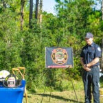 A firefighter stands next to a table with helmets and a sign displaying the number An American flag and grassy trees are in the background, under a clear blue sky