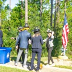 A group of people in business attire, wearing firefighter helmets, stands outdoors near an American flag and a blue table with a logo The scene is set amidst trees under a clear sky