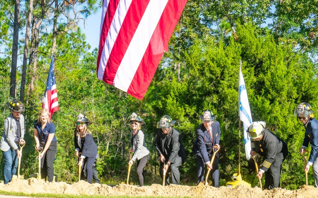 People wearing helmets and formal attire participate in a groundbreaking ceremony, shoveling dirt in a sunny outdoor setting An American flag and another flag are visible, with trees in the background