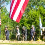 People wearing helmets and formal attire participate in a groundbreaking ceremony, shoveling dirt in a sunny outdoor setting An American flag and another flag are visible, with trees in the background