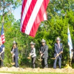 A group of people in hard hats, including firefighters, participate in a groundbreaking ceremony They hold shovels ready to dig into the soil An American flag is prominently displayed above them The background features trees and greenery