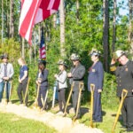 A group of people wearing hard hats and holding shovels participate in a groundbreaking ceremony They stand in a line on a dirt mound, with trees and several American flags in the background