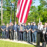 A group of people, including uniformed officials and civilians, stand in front of a large American flag outdoors Some hold a plaque Trees and greenery form the background, and a podium with a logo is in the foreground