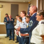 A group of military personnel stands in a line indoors, holding certificates A man in uniform claps in the background An American flag is partially visible beside them The room has beige walls and a tiled floor