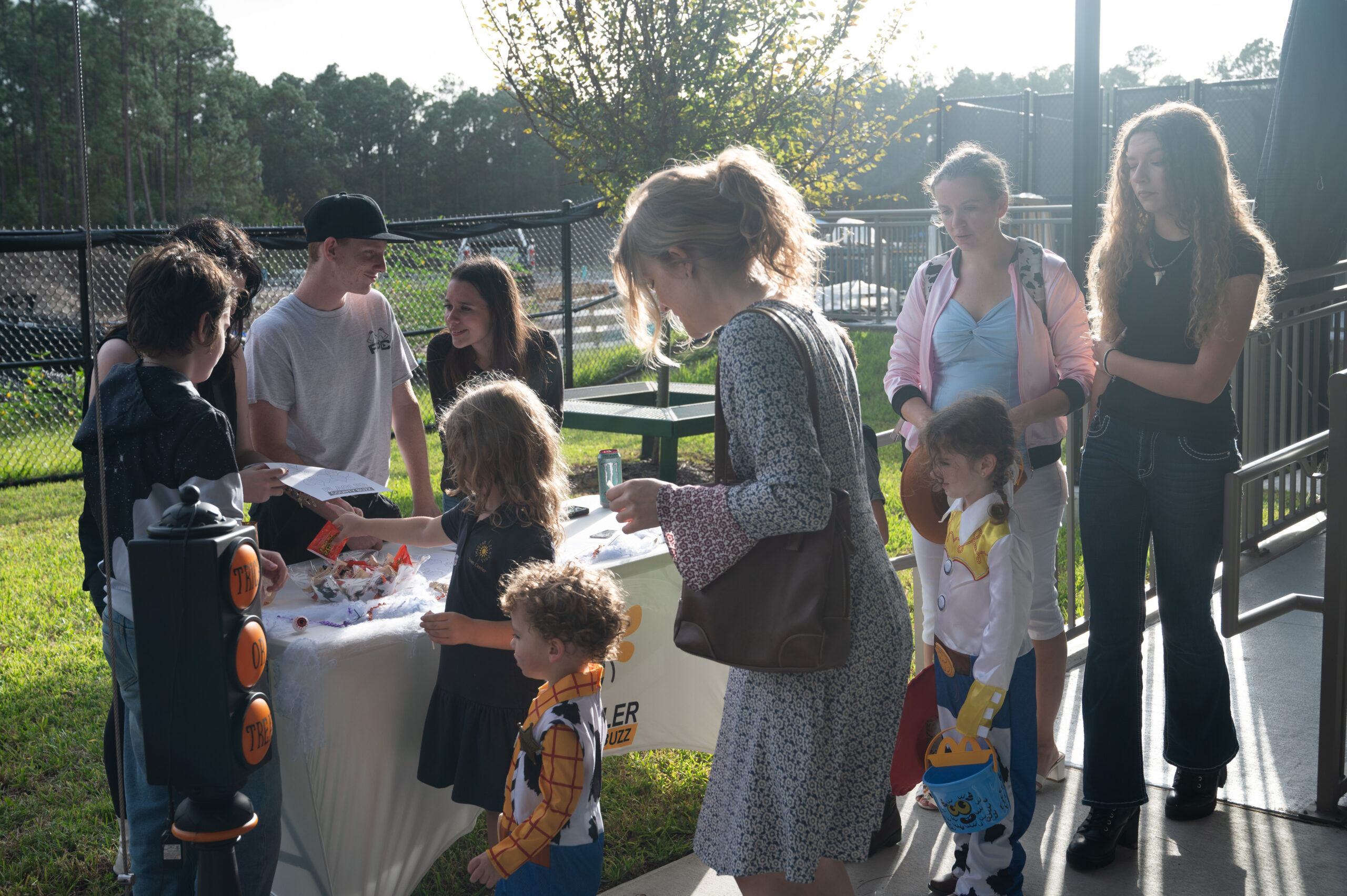 A group of people, including adults and children, gather around a table outdoors The table has items on it, with a sign visible It's a sunny day, and some children are dressed in costumes A fence and trees can be seen in the background