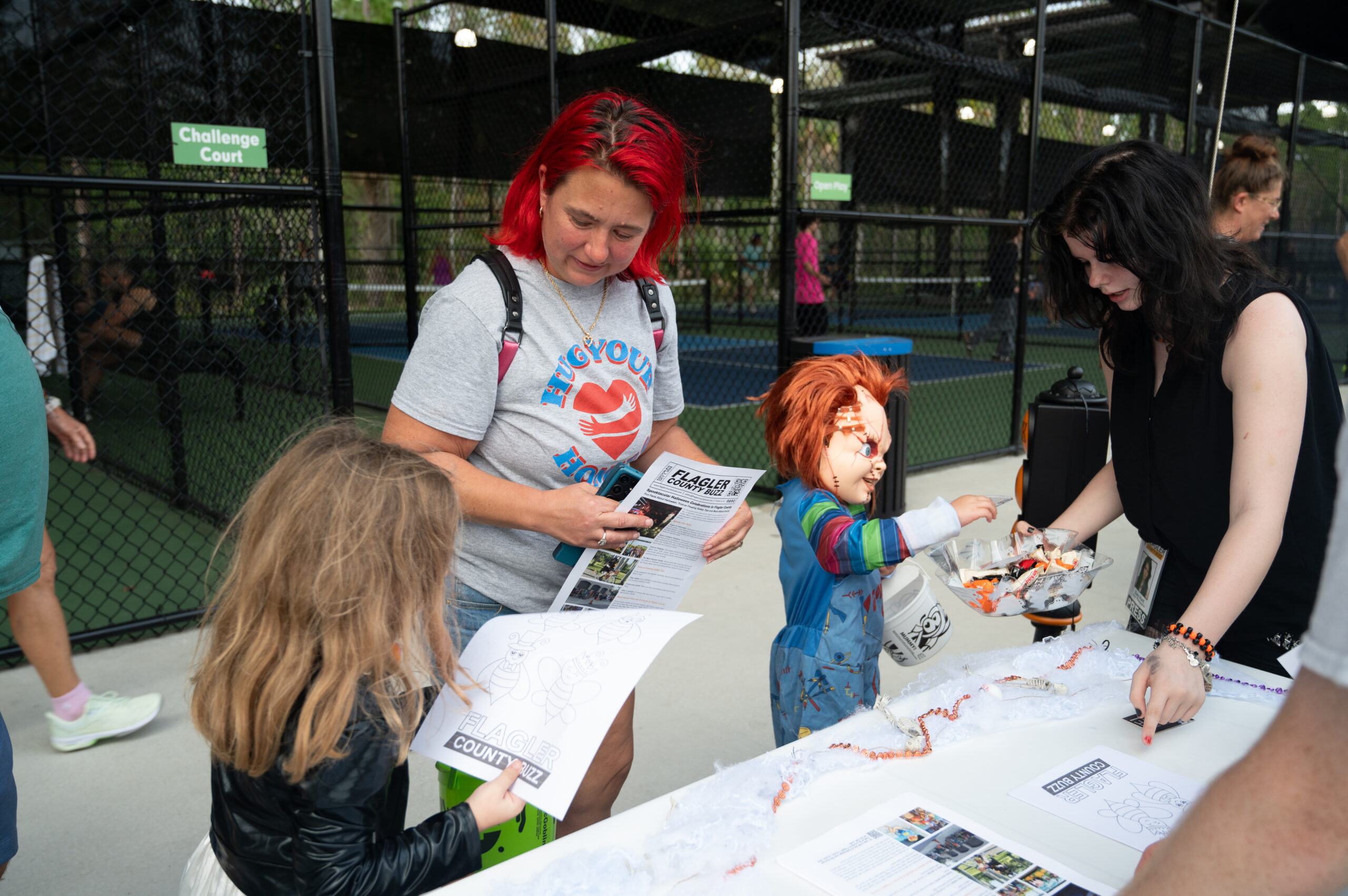 A woman and child examine papers on a table decorated for Halloween Another woman, dressed as Chucky, hands out candy They are outdoors near a sports court with a chain link fence in the background