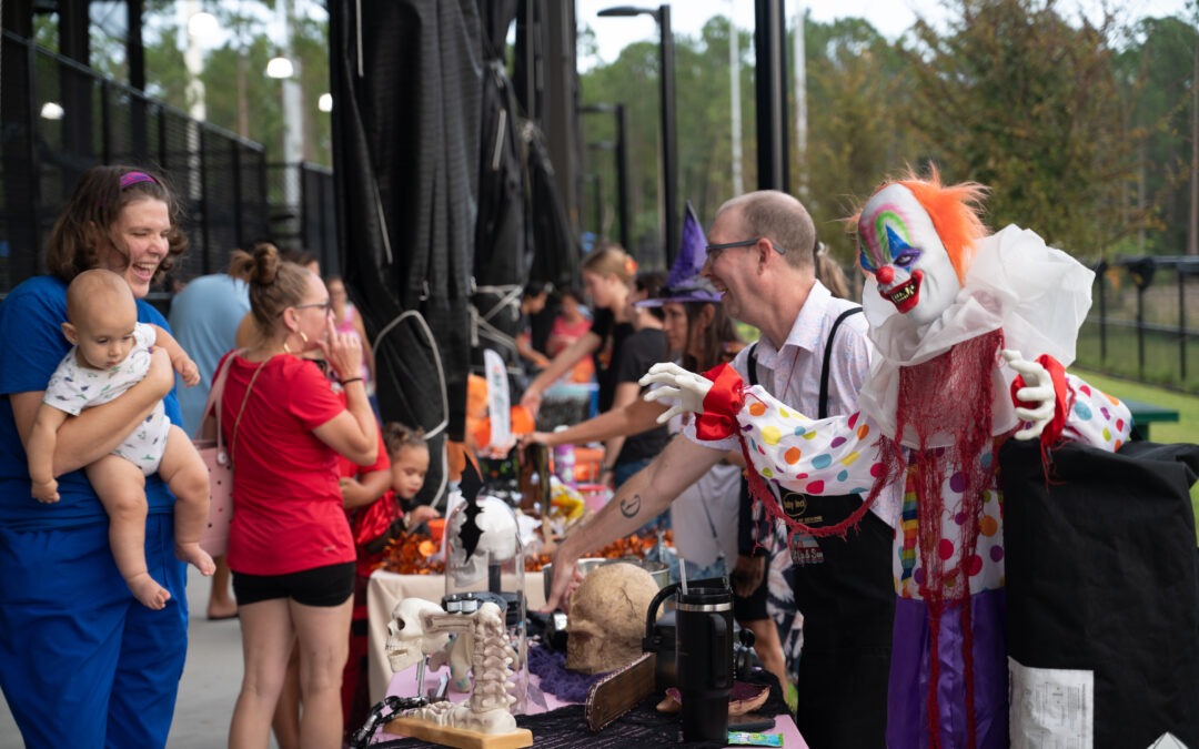 People at a Halloween themed event gather around tables with decorations A person dressed as a clown interacts with attendees while others browse items on display, including skulls and skeletons Children and adults alike enjoy the festive atmosphere