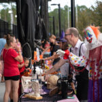 People at a Halloween themed event gather around tables with decorations A person dressed as a clown interacts with attendees while others browse items on display, including skulls and skeletons Children and adults alike enjoy the festive atmosphere