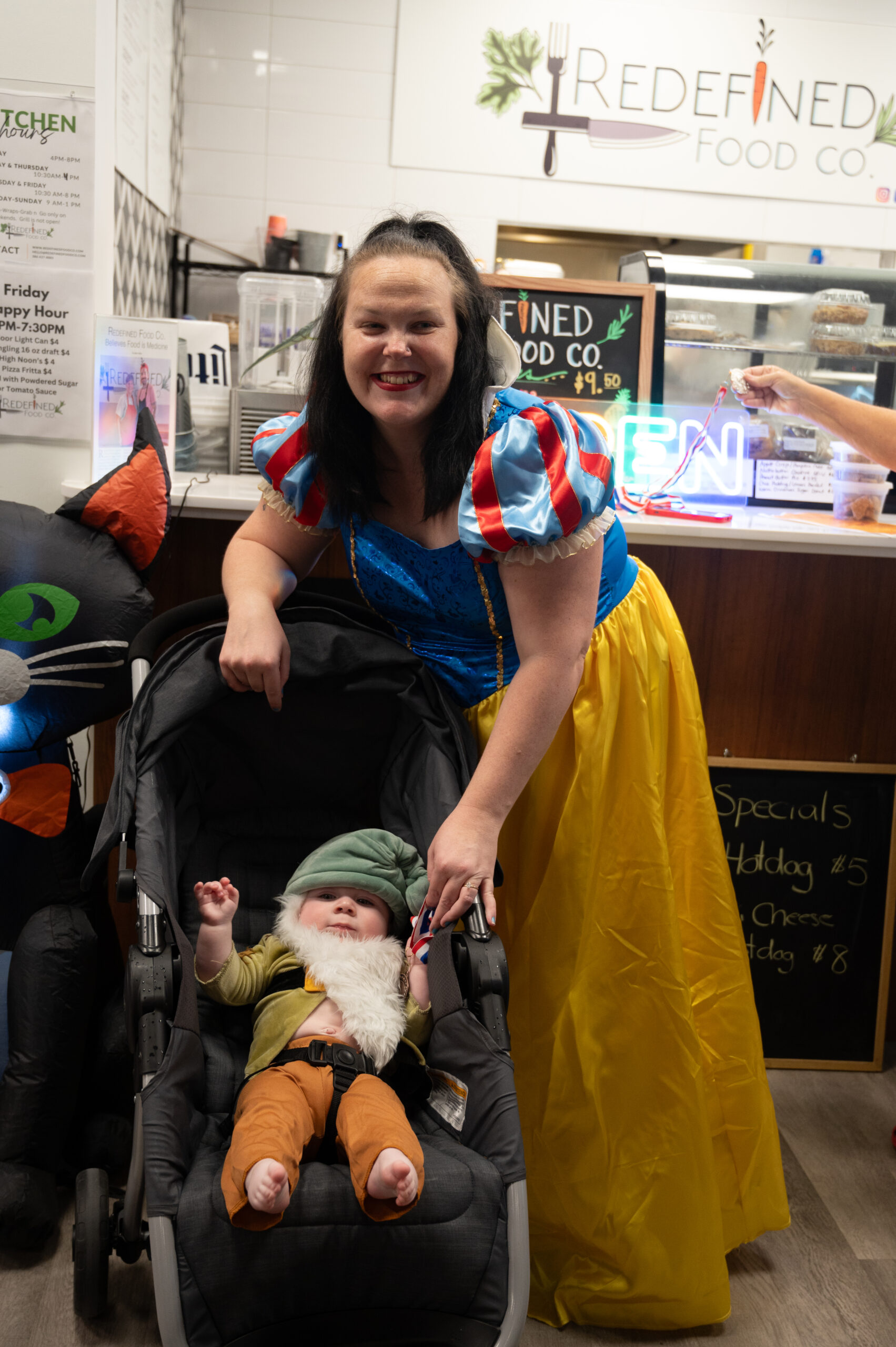 A woman dressed as Snow White smiles beside a baby in a stroller, dressed as a gnome They are inside a food establishment with a menu board and a black cat decoration visible in the background