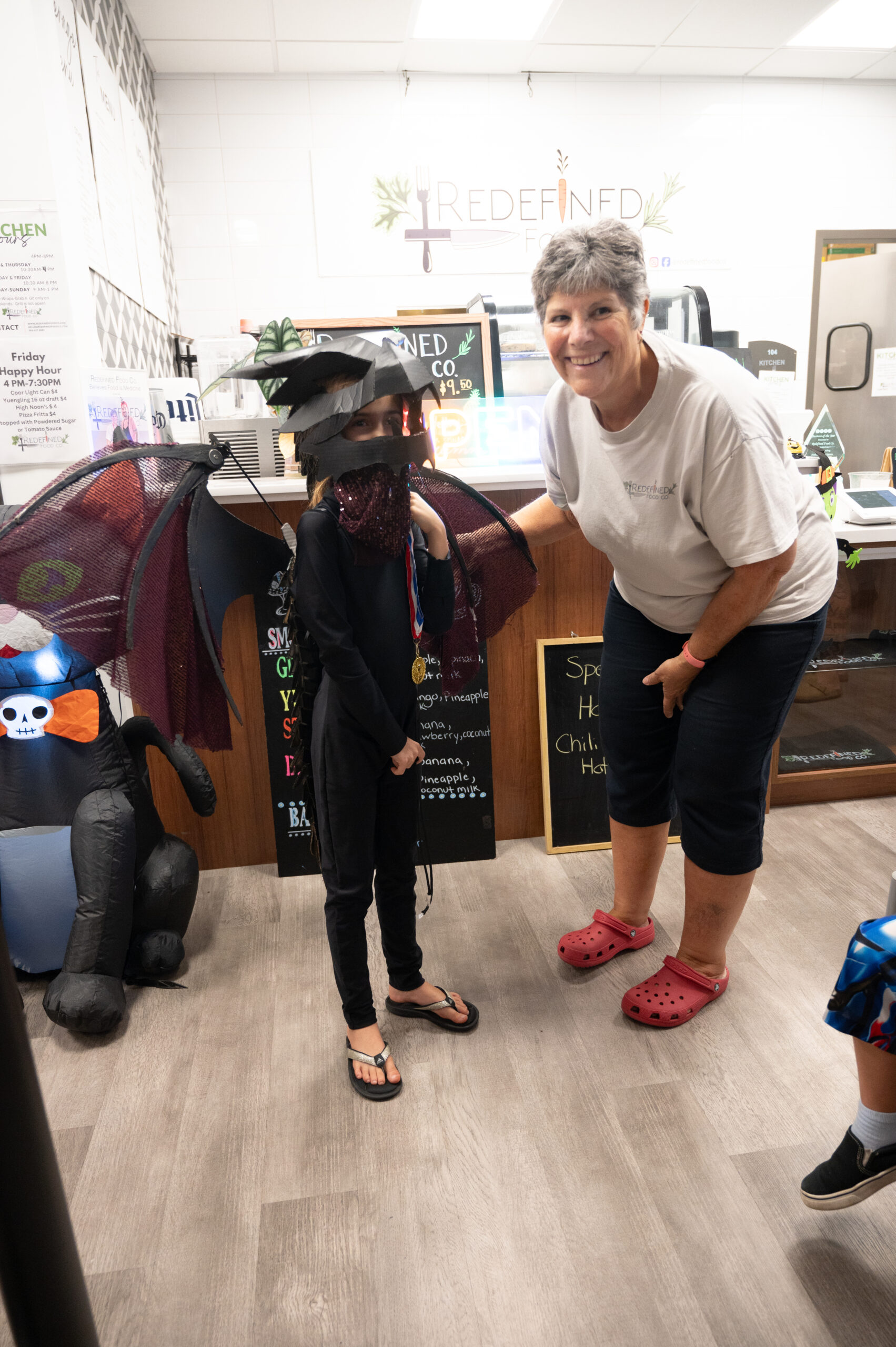 A child wearing a bat costume with wings stands next to an adult smiling at the camera The setting appears to be a store with shelves and signs in the background