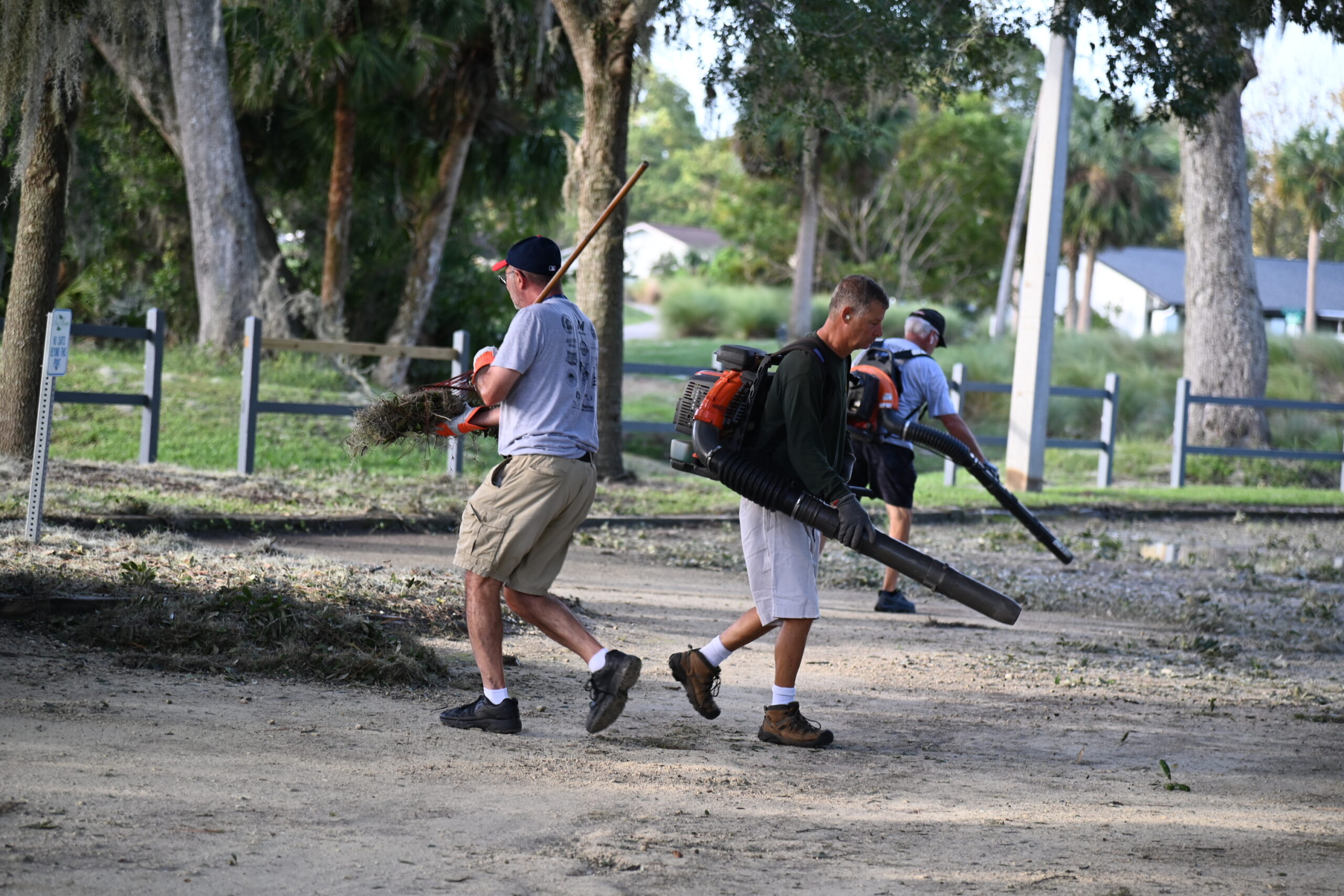 Three men are working to clean up debris in a park One carries a pile of branches, while the other two use leaf blowers Trees and a fence are visible in the background