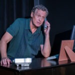 A middle aged man stands at a desk, holding a phone to his ear He wears a green polo shirt and appears engaged in conversation A computer monitor and a framed photo are on the desk in a dimly lit setting