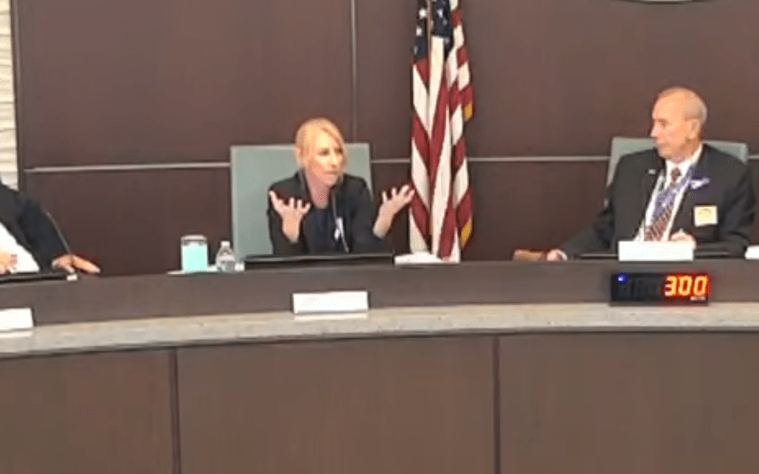 Three people are seated at a curved conference table The woman in the center is gesturing with her hands while speaking The U S flag is visible in the background A digital clock displaying ":" is on the table