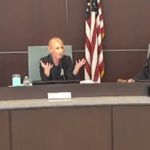 Three people are seated at a curved conference table The woman in the center is gesturing with her hands while speaking The U S flag is visible in the background A digital clock displaying ":" is on the table