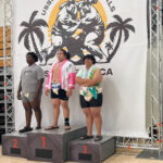 Three sumo wrestlers stand on podiums labeled , , and at the USSF Sumo Nationals A large banner displaying a sumo logo with palm trees is in the background