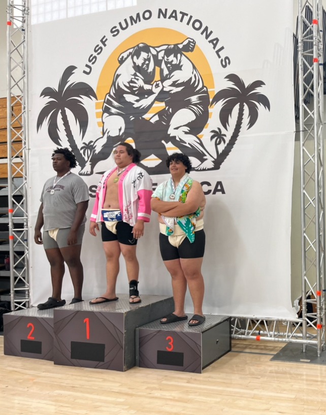 Three sumo wrestlers stand on podiums labeled , , and at the USSF Sumo Nationals A large banner displaying a sumo logo with palm trees is in the background