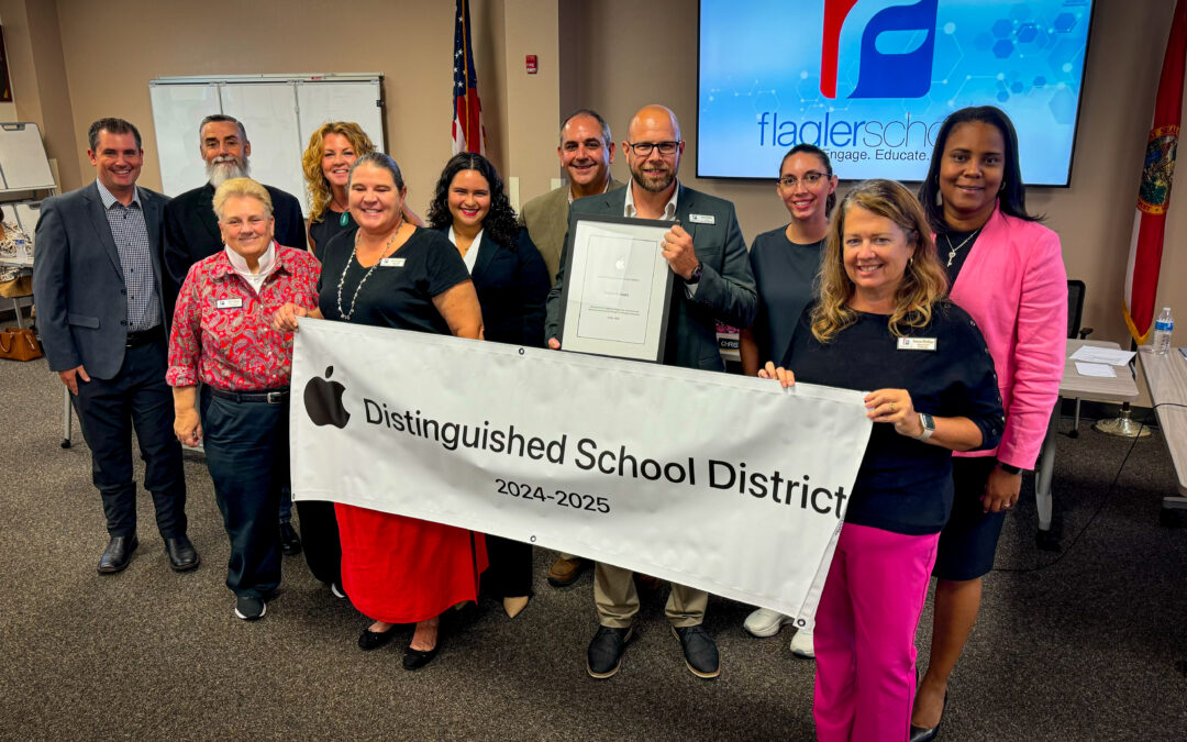 A group of people stand in a room holding a banner that reads "Apple Distinguished School District " A man in the center holds a plaque A screen displays a logo in the background