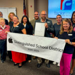 A group of people stand in a room holding a banner that reads "Apple Distinguished School District " A man in the center holds a plaque A screen displays a logo in the background