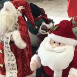 A display of Santa Claus dolls is seen, featuring one with a list of names on its coat and another with a cheerful expression wearing a red and white suit They are surrounded by festive decorations on a table indoors