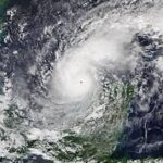 A satellite view of a swirling hurricane over the ocean The storm's eye is clearly visible at the center, surrounded by dense cloud formations The land is visible at the top and bottom edges of the image, indicating proximity to a coastline