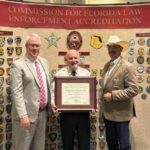 Three men stand in front of a wall filled with various law enforcement badges One man in the center is holding a certificate A banner above reads "Commission for Florida Law Enforcement Accreditation