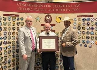 Three men stand in front of a wall filled with various law enforcement badges One man in the center is holding a certificate A banner above reads "Commission for Florida Law Enforcement Accreditation