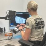 A person in a sheriff's office shirt examines a fingerprint displayed on a computer monitor They are holding an object under a camera, seated at a desk with various equipment