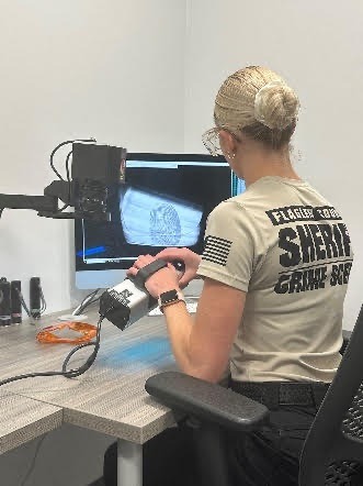 A person in a sheriff's office shirt examines a fingerprint displayed on a computer monitor They are holding an object under a camera, seated at a desk with various equipment