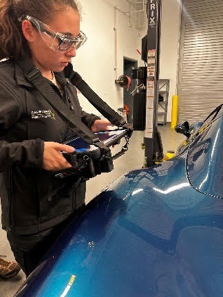 A person wearing safety goggles and a black jacket uses an augmented reality headset on a blue car's surface inside a garage Various tools and equipment are visible in the background