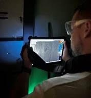 A person wearing safety glasses is holding a tablet, examining a textured surface The tablet's screen displays a grayscale image or scan The setting appears to be dimly lit, suggesting a focus on the tablet's display