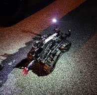 A robotic device with tank like treads is illuminated on a gravel surface at night It has a mounted light and a mechanical arm extending forward